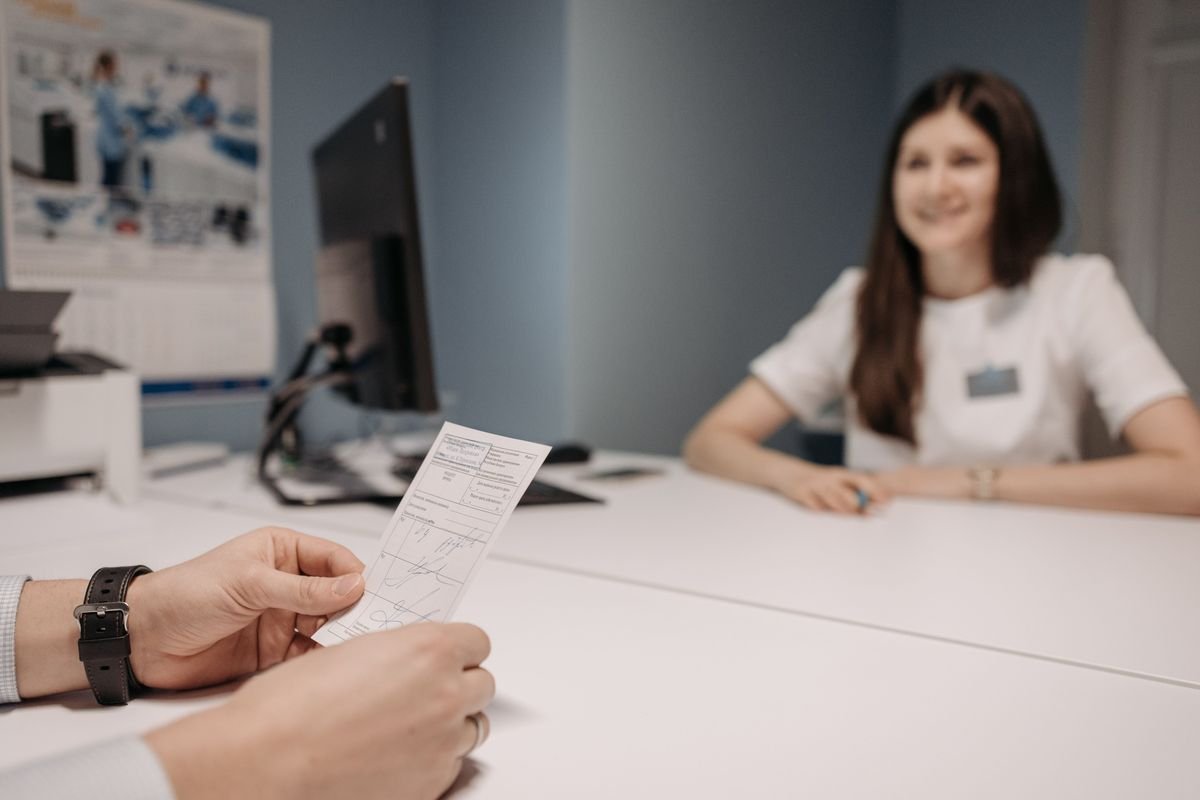 A patient's hands holding a handwritten prescription slip across a white desk from a smiling clinician in a white top.