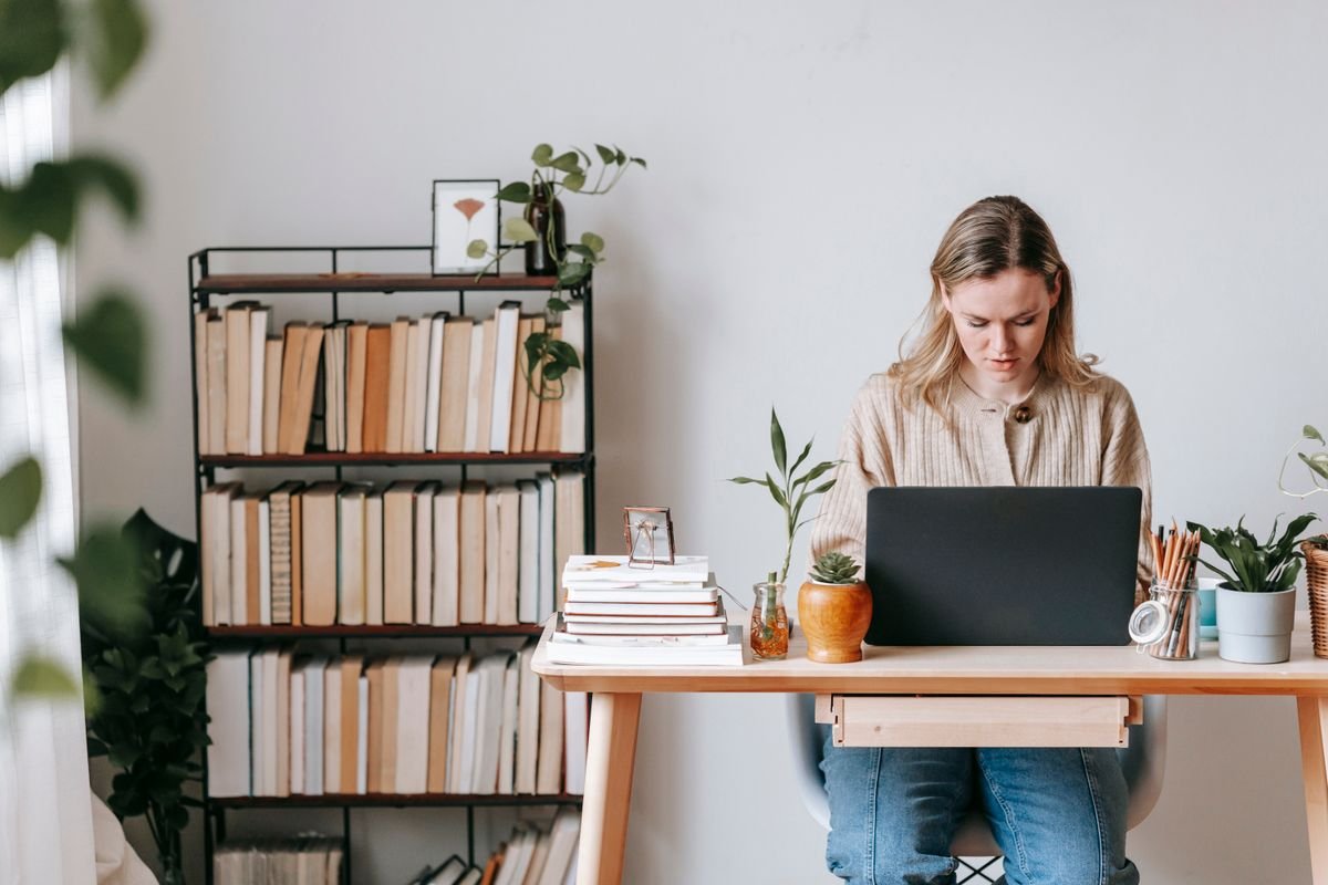 Woman working on a laptop at a home desk with potted plants and a bookcase behind her.