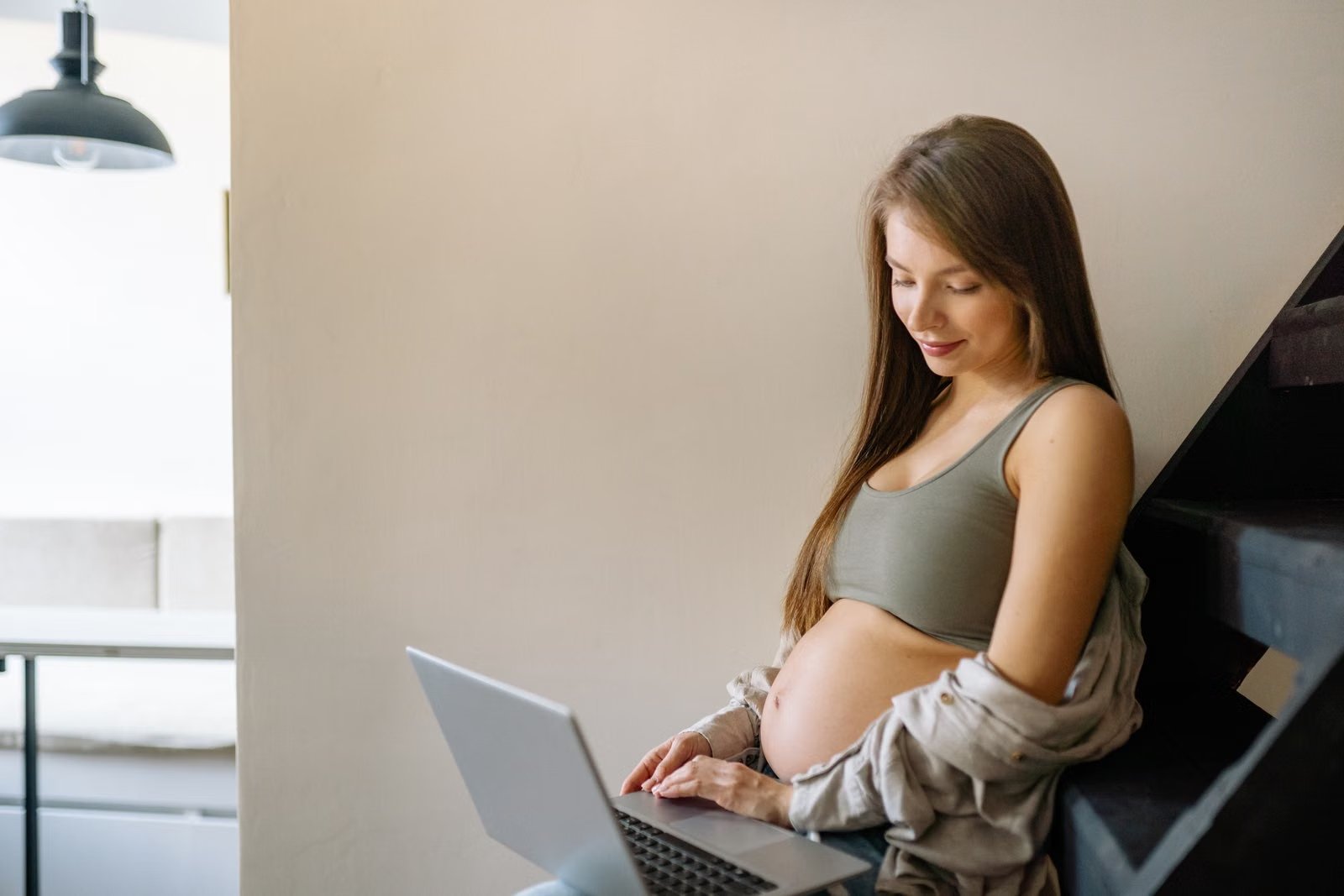 Pregnant woman sitting on a staircase working on a laptop at home.