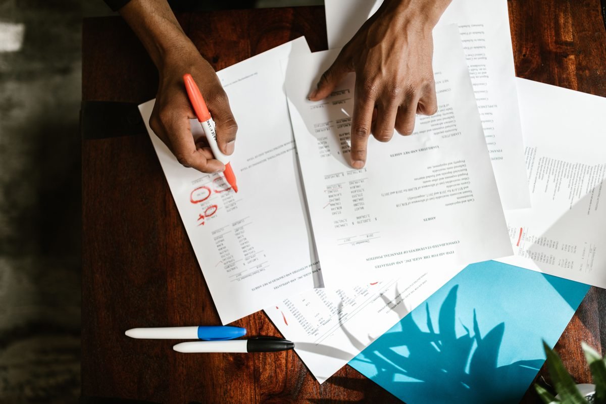 Overhead view of two people comparing printed benefit documents on a wooden desk, one marking figures with an orange pen.