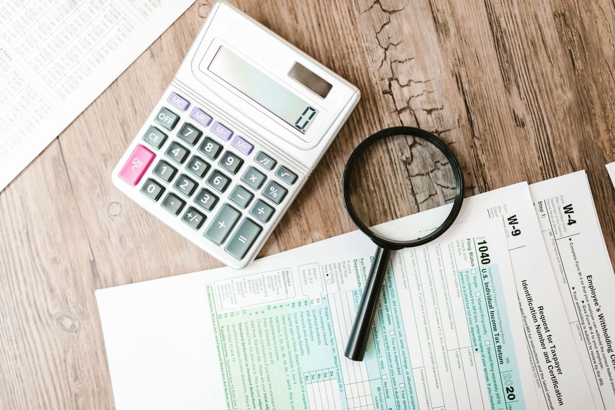 Overhead view of a calculator and a magnifying glass resting on U.S. tax forms including Form 1040 and Form W-4 on a wooden desk.